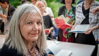 Die Autorin Helga Schubert: Links im Bild eine ältere grauhaarige weiße Frau mit strähnigem schulterlangem grauem Haar. Sie lächelt in die Kamera, im Hintergrund unscharf stehen Menschen an einem Tisch an, um sich Bücher signieren zu lassen.
