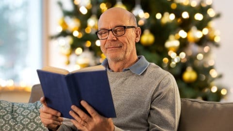 Älterer Herr mit Glatze sitzt mit einem Buch in der Hand vor dem Weihnachtsbaum.