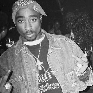 Der Rapper und Schauspieler Tupac Shakur: schwarz-weiß Bild eines afroamerikanischen Mannes in Jeansjacke und Adidas-Pullover. Er zeigt beide Mittelfinger in die Kamera. Er trägt auf dem Kopf ein Bandana und hat einen Oberlippenbart.