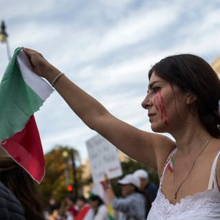 Demonstrantin hält Iranflagge in die Höhe, an den Augen ist Blut gemalt