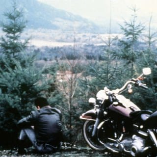 Ein Bild aus der ersten Staffel Twin Peaks zeigt einen jungen Mann, der neben seinem Motorrad sitzt und in ein Tal voller Nadelbäume blickt