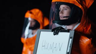 Amy Addams in einem Schutzanzug, sie hält ein Schild mit der Aufschrift "Human" in der Hand, ein Still aus "Arrival" (2016)