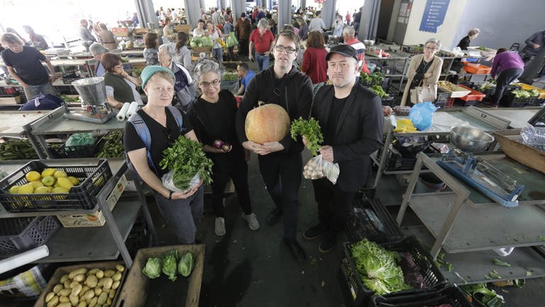 Mitglieder des Wiener Gemüseorchesters stehen mit Petersilie, Zwiebeln, Kürbis vor einem Marktstand