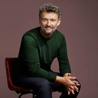 Der Tenor Jonas Kaufmann: Mann mit grau meliertem Haar sitzt mit gefalteten Händen auf einem Stuhl und schaut in die Kamera.