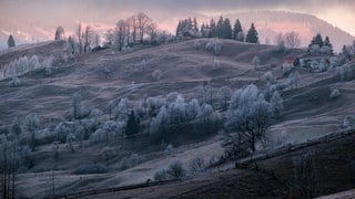 Auf dem Bild ist ein Dorf mit Hügeln  bei Sonnenuntergang zu sehen. Auf den Bäumen ist Raureif.