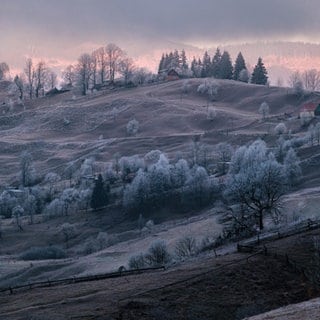 Auf dem Bild ist ein Dorf mit Hügeln  bei Sonnenuntergang zu sehen. Auf den Bäumen ist Raureif.