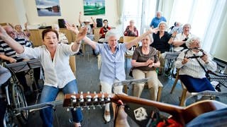 Blick über die Schulter eines Gitrarristen: Senior*innen singen im Sitzen, einige haben Intrumente wie einen Schellenkranz oder Perkussionsinstrumente. Andere strecken freudig die Hände in den Himmel.