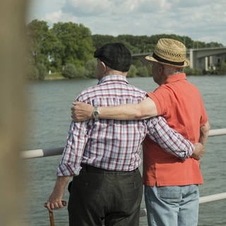 Symbolbild zum Tag der Freundschaft: Zwei ältere Männer stehen Arm in Arm an einem Flussufer