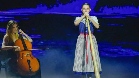 Alphornistin Melina Grimm spielt in einem blauen Dirndl das Alphorn. Links neben ihr sitzt eine Cellistin.