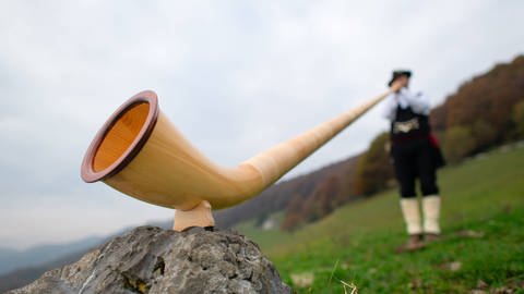 Alphorn auf einer Alpenwiese. Ein Mann in Tracht ist im Hintergrund zu sehen, wie er das Alphorn spielt.