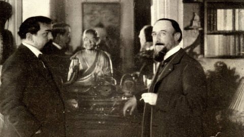 Fotografie der Komponisten Claude Debussy und Erik Satie, beide rauchend an einen Kamin gelehnt. Auf dem Kaminsims thront eine Buddha-Statue.