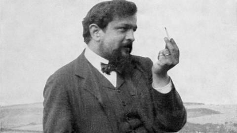 Schwarz-Weiß-Fotografie von Claude Debussy. Er lehnt auf die Ballustrade eines Balkons und sieht liebevoll zu seiner Zigarette, die er erhoben hält.