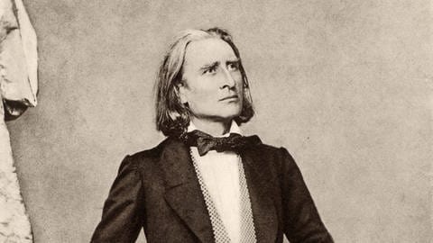 Schwarz-Weiß-Porträt des jungen Franz Liszt. Der Komponist steht in einem Anzug aufrecht, den Arm auf eine Stuhllehne angelegt.