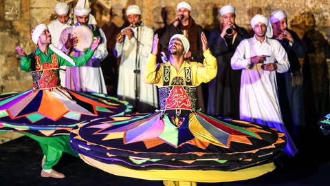 Zwei Männer in bunten Tellerröcken tanzen die Tanoura. Im Hintergrund stehen Musiker in schwarzen und weißen Kaftans.