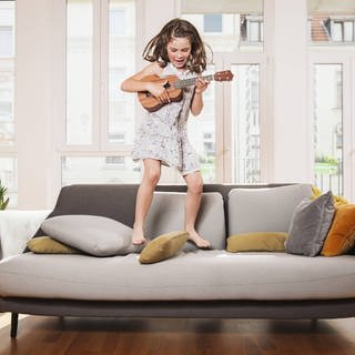 Symbolbild Musik und Bewegung: Ein Mädchen spielt Ukulele und springt auf einem Sofa