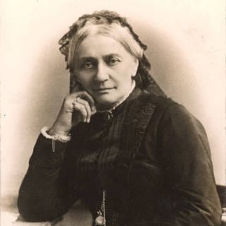 Fotografie der betagten Clara Schumann von Wilhelm Dreesen (1899). Schumann blickt direkt in die Kamera, das Kinn leicht auf die rechte Hand gestützt. Sie trägt ein schwarzes Kleid und eine schwarze Haube.