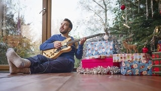 Ein Mann sitzt vor einem Weihnachtsbaum und musiziert mit einer E-Gitarre