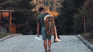 Mann läuft mit Tuba einen Weg entlang, Ansicht von hinten