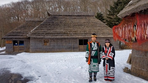 Ein Mann und eine Frau in der Tracht der Ainu posieren im Ainu-Museum von Shiraoi vor traditionellen Ainu-Häusern