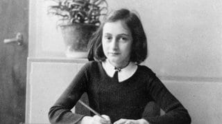 Anne Frank (1929-1945) sitzt an einem Tisch. Sie schreibt mit einem Bleistift in ein Heft.