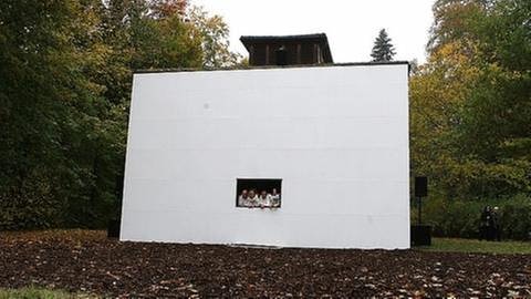 Riesige weiße Bühnenwand mit Fenster am Donaueschinger Fischerhaus, aus dem vier Leute rauskucken