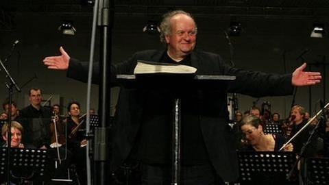 Komponist Wolfgang Rihm seht auf dem Dirigentenpotest und breitet die Arme zum Publikum aus