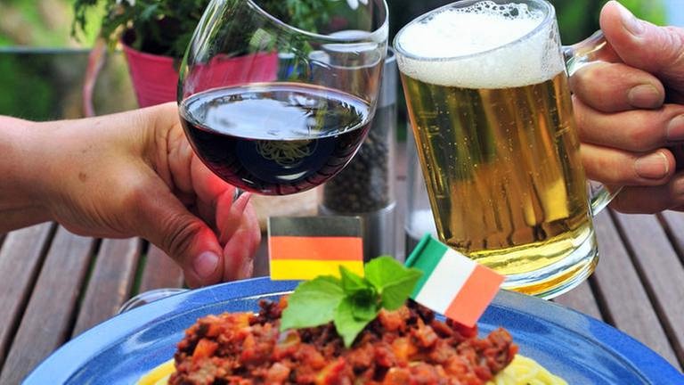Mit italienischem Rotwein und deutschem Bier stoßen ein Mann und eine Frau an. Im Vordergrund steht ein Teller Spaghetti, geschmückt mit einer deutschen und einer italienischen Flagge.