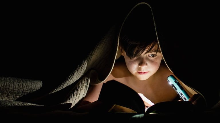 Ein Kind liegt im Bett und liest unter der Decke ein Buch bei Nacht