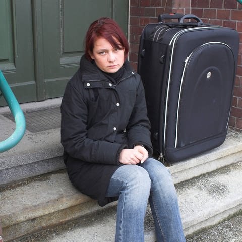 Junge Frau sitzt allein mit Koffer auf einer Treppe: Mit 18 Jahren erwachsen – für Pflege- und Heimkinder endet dann in der Regel die Jugendhilfe. Die jungen Menschen stehen plötzlich alleine da und werden zu Niemandskindern: Niemand fühlt sich für sie verantwortlich – auch die Behörden nicht.