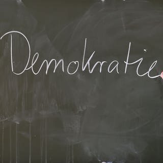 Ein Lehrer schreibt mit Kreide das Wort "Demokratie" an eine Schultafel: Kinder und Jugendliche sollten möglichst früh ihre Rechte, Pflichten und ihren Selbstwert in der Gesellschaft kennen: teilhaben, mitgestalten und Eigenverantwortung übernehmen von Anfang an