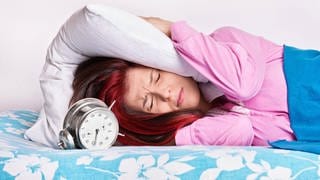 Frau hält sich Kissen aufs Ohr, um den Wecker nicht zu hören: Jeder sollte so lange schlafen, bis er ausgeschlafen ist. Wer zu lange im Bett bleibt oder zu kurz schläft, kann Schlafstörungen entwickeln.