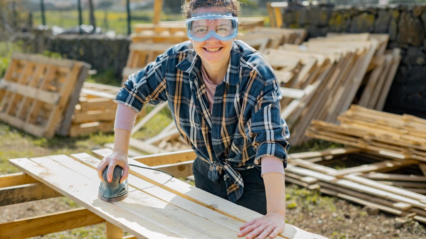 Frau mit Schutzbrille schleift Bretter auf einer Baustelle: Ob auf dem Dachstuhl, der Malerleiter oder am Sicherungskasten – Handwerkerinnen sind immer noch die große Ausnahme.