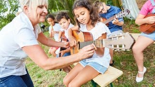 Kinder lernen Gitarre spielen, eine Lehrerin hilft: Durch Musik lernen Kinder und Jugendliche, einander zuzuhören, erleben Gemeinschaft und verstehen die Kultur eines Landes. Dennoch fällt Musikunterricht regelmäßig aus oder wird fachfremd unterrichtet. 