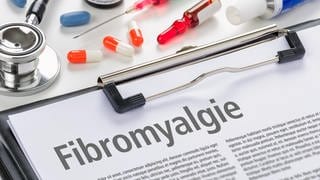Klemmbrett mit einem Infoblatt zur Fibromyalgie, dazu Medikamente und ärztliche Instrumente:Wer an Fibromyalgie leidet, hat oft starke Schmerzen. Meist trifft es Frauen. Lange galt die Erkrankung als psychosomatisch. Dabei sind die Ursachen komplex – und noch unklar.