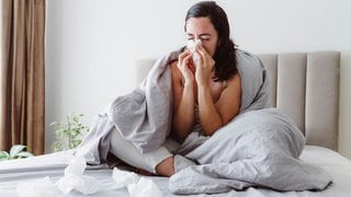 Eine Frau sitzt krank auf dem Bett und putzt sich die Nase.