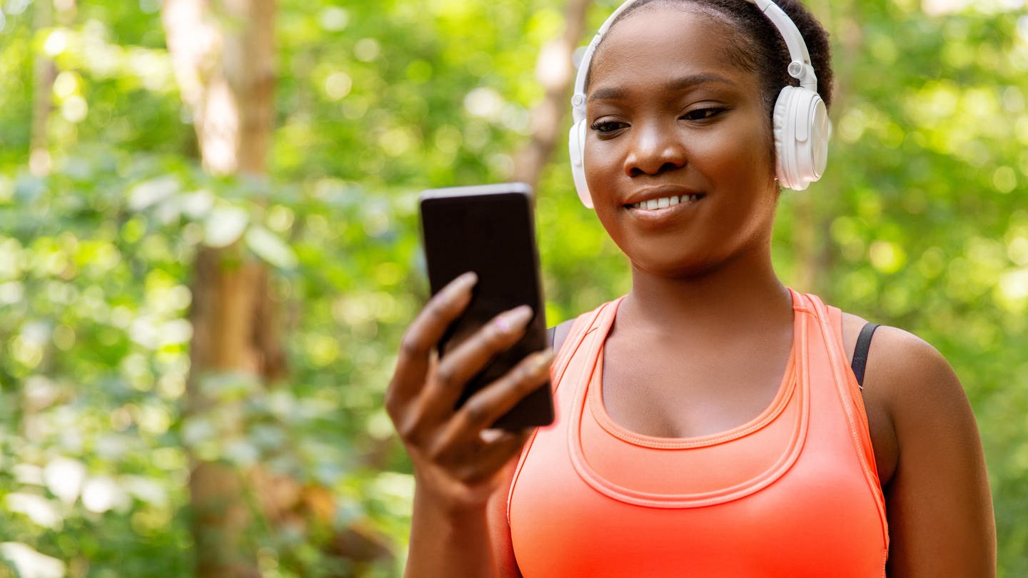 Junge Frau mit Kopfhörer und Smartphone geht durch den Wald: Das Radio der Zukunft soll die Höererinnen und Hörer unterwegs mit relevanten Informationen versorgen und mittel KI mit ihnen in Dialog treten können.