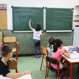 Klassenzimmer einer italienischen Grundschule. Ein kleines Mädchen schreibt auf die Tafel "viva la scuola", Es lebe die Schule.