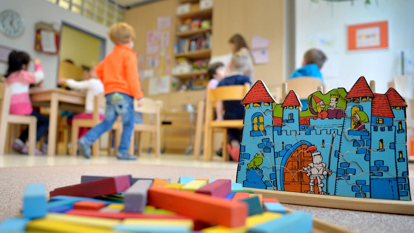 Spielzeug liegt in einer Kindertagesstätte auf dem Boden, im Hintergrund Kinder.
