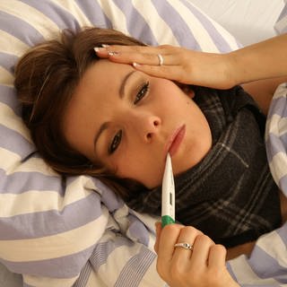Eine junge Frau liegt mit Fieberthermometer im Bett.