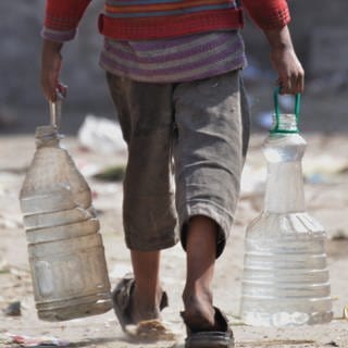Ein Mann trägt in beiden Händen große FlaschenKanister voller Wasser. 