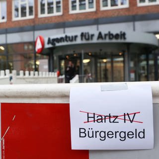 Ein Blatt Papier mit dem Wörtern Harz IV (rot durchkreuzt) und Bürgergeld ist vor einer Baustelle der Agentur für Arbeit zu sehen. Der Bundestag hat für die Einführung des Bürgergelds gestimmt. Es löst 2023 die bisherigen Leistungen von Hartz-IV ab.