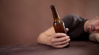 Ein Mann liegt mit dem Oberkörper an der Tischkante und hält eine Flasche Bier in der Hand