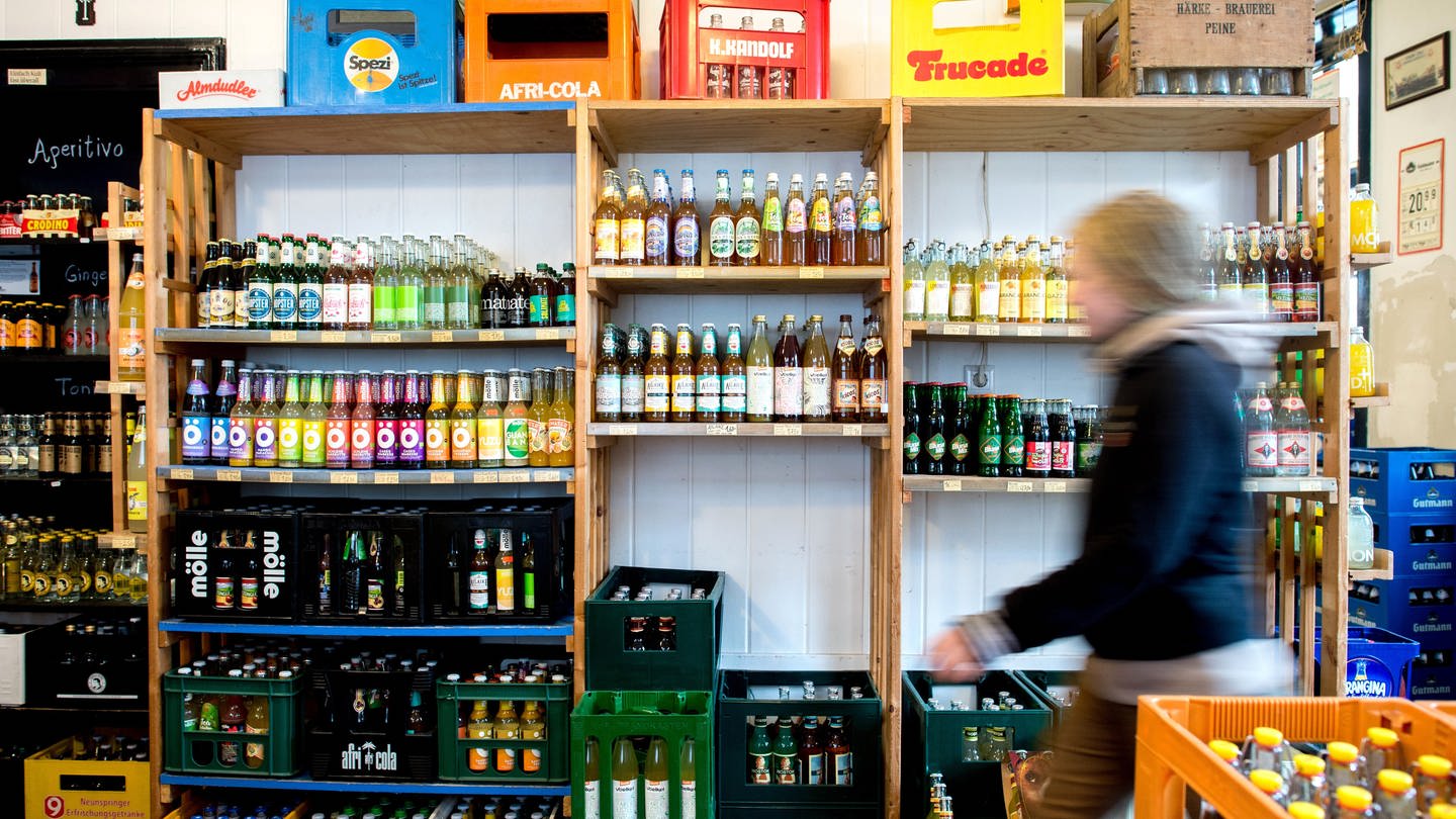 Eine Frau geht in einem Getränkemarkt an einem Regal vorbei, in dem Limonaden verschiedener Marken stehen.