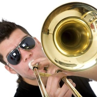 Junger Mann mit Sonnenbrille bläst in eine Posaune: Die Wucht von Trompete, Posaune und Tuba, moderne Rhythmen und Elemente von Techno bis Pop – neue Blasmusik-Combos sind mitreißend.