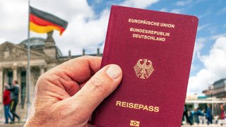 Hand eines Mannes hält deutschen Reisepass, im Hintergrund der Berliner Reichstag mit Deutschlandfahne: Seit Juni 2024 erleichtert in Deutschland ein Gesetz die Einbürgerung. Kritiker sagen, die Voraussetzungen seien nicht streng genug. Befürworter sehen durch Teilhabe die Demokratie gestärkt. 