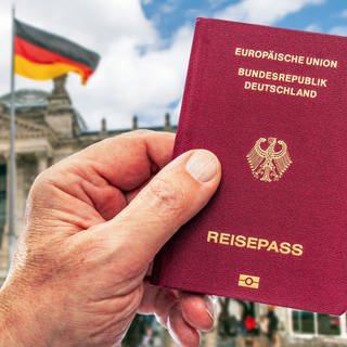 Hand eines Mannes hält deutschen Reisepass, im Hintergrund der Berliner Reichstag mit Deutschlandfahne: Seit Juni 2024 erleichtert in Deutschland ein Gesetz die Einbürgerung. Kritiker sagen, die Voraussetzungen seien nicht streng genug. Befürworter sehen durch Teilhabe die Demokratie gestärkt. 