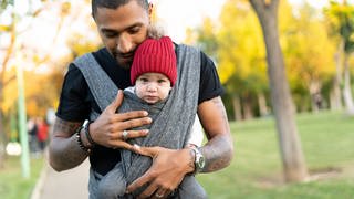 Vater hält schützend seine Hande um das Baby, das er im Tragetuch vor sich hält: Heute möchten immer mehr junge Männer liebevolle und präsente Väter sein. Und auch die Forschung ergründet immer besser, was Männer im Umgang mit Kindern oft anders machen. Klar ist: Kinder können in ihrer Entwicklung sehr von aktiven Vätern profitieren.