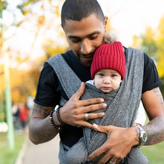 Vater hält schützend seine Hande um das Baby, das er im Tragetuch vor sich hält: Heute möchten immer mehr junge Männer liebevolle und präsente Väter sein. Und auch die Forschung ergründet immer besser, was Männer im Umgang mit Kindern oft anders machen. Klar ist: Kinder können in ihrer Entwicklung sehr von aktiven Vätern profitieren.