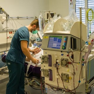 Ein Pfleger kümmert sich um einen Patienten, der während seines Aufenthalt in einem Intensivzimmer an einem Beatmungsgerät und einem Dialysegerät hängt.