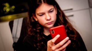 Ein Mädchen schaut ernst auf sein rotes Smartphone: Immer mehr Teenager verlieren sich in der digitalen Welt und kämpfen mit der Realität, einige entwickeln Suchtverhalten, Ängste oder Depressionen. Medizin wie Wissenschaft entwickeln für die wachsende Zahl Betroffener kreative Therapien und präventive Ansätze und holen die Eltern mit ins Boot.